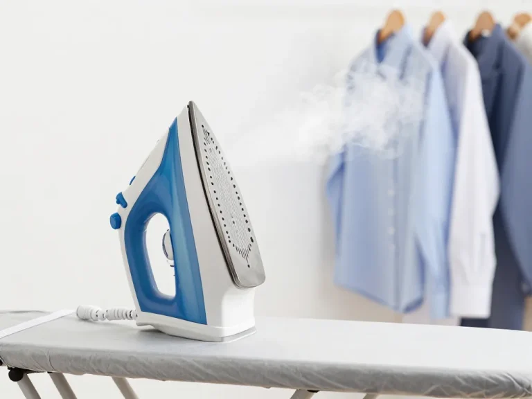 A high-end steam iron on an ironing board, with a powerful jet of steam rising, against a clean white background. The iron has a metallic ceramic soleplate visible, a water tank, and a cable. The scene looks professional, clean, and modern, emphasizing efficiency and home use. In the background, neatly stacked shirts and dresses on hangers.
